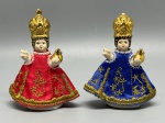 ARTE SACRA - Lote de 2 esculturas, representando Menino Jesus de Praga, confeccionado em porcelana com acabamento esmaltado, coroas e detalhes dourados, e vestimentas em tecido acetinado (vermelho e azul) com bordados em fio dourado. Aureta Figural, República Tcheca (conforme marcação na base). Alt. 8 cm. Pequenas faltas e cabeças coladas.
