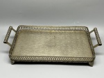 METAL - Elegante bandeja retangular em ferro espessurado a prata. A peça destaca-se pelo fundo lavrado com delicado padrão texturizado, galeria delicadamente recortada nas bordas, pés estilizados e alças vazadas. Med. 8x37x20 cm. Marcas do tempo e de uso.