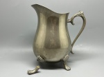 METAL - Elegante jarra antiga em metal prateado, com corpo bojudo, alça lateral trabalhada com detalhe floral e bico vertedor proeminente. A peça apoia-se sobre pés ornamentados em formato de garra com detalhes de vieira (nota-se pé solto/descolado que requer restauro). Alt. 21 cm. Marcas do tempo e de uso.