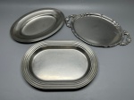 INOX - Conjunto composto por três travessas ovais em aço inoxidável com diferentes designs: uma com bordas limpas e clássicas, uma com belas alças laterais trabalhadas e bordas ornamentadas em relevo, e uma terceira com detalhe de frisos concêntricos em relevo na aba. Med. 38x23 cm, 35x25 cm e 31x22 cm. Marcas do tempo e de uso.