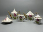 PORCELANA - Elegante conjunto em porcelana branca composto por peças de design clássico com detalhes e pinturas douradas nas alças, bicos e bordas, apresentando uma decoração com delicados arranjos florais de rosas cor-de-rosa, botões e folhagens. O lote integra bule de chá com tampa, bule de café esguio com bico e alça dourados, leiteira de formato cônico, açucareiro com tampa e pegador cônico, além de uma manteigueira ou peça funda com prato de base e tampa cônica decorada. Maior 19 cm e menor 10 cm.