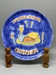 CERÂMICA - Prato decorativo de cerâmica esmaltada com fundo azul, apresentando pintura figurativa central que retrata uma ave vestida de chef de cozinha sobre uma travessa, flanqueada por talheres e taças, com as inscrições pintadas "Cantina Paludo" na parte superior e "Niterói - RJ" na parte inferior. O verso possui acabamento esmaltado com craquelê característico, perfuração própria para fixação na parede e assinatura manuscrita da ceramista "Liliane Porto" com monograma. Med. 23 cm. Marcas do tempo e de uso.