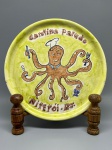 CERÂMICA - Prato decorativo de cerâmica esmaltada com fundo em tons esverdeados, apresentando pintura figurativa central que retrata um polvo antropomórfico vestido de chef de cozinha (com chapéu e gola), segurando talheres, taça e guardanapo em seus tentáculos. O conjunto conta com as inscrições em relevo/pintadas "Cantina Paludo" na parte superior e "Niterói - RJ" na parte inferior. O verso exibe acabamento esmaltado com craquelê característico, suporte integrado para fixação em parede e a assinatura manuscrita da ceramista "Liliane Porto" com monograma. Dia. 23 cm. Marcas do tempo e de uso.