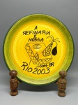 CERÂMICA - Prato decorativo em cerâmica artesanal esmaltada, apresentando fundo em tons de amarelo e borda verde. O centro exibe pintura temática alusiva à indústria petrolífera, com a representação de uma plataforma/torre de extração e a inscrição em destaque: "A REFINARIA É NOSSA", acompanhada dos dizeres "Rio 2003" e indicação de site. O verso possui carimbo e marcação manuscrita "HAND PAINTED / bric - Dia. 25 cm.