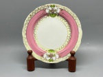 PORCELANA - Prato decorativo em porcelana fina de fabricação alemã, apresentando borda com acabamento ondulado e recortes geométricos em relevo. A aba é composta por uma marcante faixa em tom rosa e detalhe interno em amarelo claro com motivos geométricos zigue-zague. O destaque decorativo fica por conta de medalhões opostos com detalhes em relevo texturizado em verde e aplicações florais pintadas à mão com toques em dourado. O verso exibe a marca de fabricação "Oskar Schaller / Kirchenlamitz / Bavaria" encimada por coroa e monograma, além da numeração 101. Dia. 19 cm. Marcas do tempo e de uso.