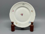 PORCELANA - Prato decorativo em porcelana branca de fabricação fina, com aba de borda ondulada adornada por relevo texturizado em motivos vegetal/geométricos contínuos e filete dourado na extremidade. O espelho apresenta um delicado bouquet floral policromado ao centro, complementado por pequenos raminhos de flores coloridas dispersos pela aba. O verso exibe marca de fabricação com o emblema de águia encimada por letras e a inscrição "CZECHOSLOVAKIA", além de marcação numérica de época. Dia. 17 cm. Marcas do tempo e de uso.