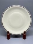 PORCELANA BAVARIA - Prato raso em cerâmica ou porcelana com aba lisa, destacado por uma elegante faixa decorativa perimetral em tom preto com padrão geométrico e estilizado de inspiração clássica, finalizado com um delicado filete dourado na borda. O espelho apresenta superfície lisa em tom off-white. Dia. 24 cm. Marcas do tempo e de uso.