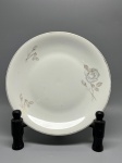 PORCELANA - Prato raso em porcelana branca de fabricação nacional, com aba lisa decorada com delicados motivos de rosas e botões em tons monocromáticos acinzentados, acompanhados de folhagens e acabamento com filete dourado na borda. O verso apresenta o selo de fabricação em verde com a inscrição "PORCELANA RENNER PORTO ALEGRE". Dia. 25 cm. Marcas do tempo e de uso.