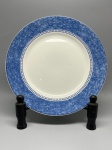 PORCELANA - Prato raso em cerâmica ou porcelana com aba larga decorada em padrão texturizado azul em efeito marmorizado ou esponjado, delimitada internamente por uma faixa geométrica contínua em formato de pequenos quadrados (grega estilizada) sobre fundo branco. O espelho apresenta superfície lisa em tom off-white. O verso exibe o selo com a inscrição cursiva "Normandie" ornamentada por motivos florais estilizados. Dia. 26,5 cm. Marcas do tempo e de uso.