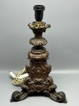 ABAJUR - Antiga base de abajur em metal trabalhado com rica ornamentação em relevo de volutas e motivos clássicos, estruturada sobre base tripé, com altura de 38 cm, apresentando desgastes naturais do tempo e marcas de uso.