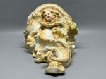 BISCUIT - Peanha (ou modilhão) de parede em biscuit europeu, característica do estilo Art Nouveau, ornamentada com a face de um querubim e detalhes em relevo com acabamento dourado, medindo 20x15x13 cm e apresentando desgastes naturais do tempo, pequenas bicaduras e marcas de uso.