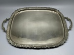 METAL - Bandeja retangular de cantos arredondados, em metal espessurado a prata, apresentando borda decorada com trabalho em relevo de cachos de uvas e folhagens, acompanhada por alças laterais com o mesmo padrão ornamental. Medindo 60x43 cm, a peça apresenta desgastes naturais do tempo e marcas de uso.