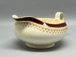 FAIANÇA - Molheira em faiança inglesa da marca Alfred Meakin, apresentando design clássico com bico vertedor proeminente, alça lateral ergonômica, base circular elevada e fundo creme. Decorada com faixa superior em tom bordô arrematada por friso dourado e delicados motivos estilizados repetidos ao longo do corpo. Peça marcada no verso, ostentando o clássico efeito craquelê da pasta cerâmica, além de desgastes naturais do tempo, pequenas marcas de uso e oxidação.