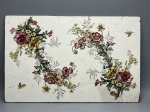 FAIANÇA - Placa decorativa antiga em faiança, decorada com delicados arranjos florais policromados destacando flores em tons de vinho e amarelo (como papoulas e margaridas), folhagens detalhadas e pequenas borboletas pintadas nos cantos. A peça apresenta fundo esmaltado em tom off-white com sutis salpicados característicos da época, verso liso esmaltado e desgastes naturais nas bordas compatíveis com sua antiguidade.