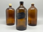 DEMI CRISTAL - Lote com três garrafas farmacêuticas de vidro âmbar com 23 cm de altura cada, sendo uma delas rotulada e todas acompanhadas de tampas.