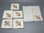 CAMA - MESA - BANHO - Lote de 18 peças de enxoval vintage em tecido claro, composto por guardanapos e peças de mesa bordadas à mão em ponto cruz com motivos florais coloridos e acabamento em caseado azul nas bordas, apresentando marcas do tempo e manchas amareladas.