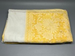 CAMA - MESA - BANHO - Toalha de mesa (300x170 cm) em tecido misto combinando painéis em tecido liso claro com faixas em tom amarelo decoradas com grandes motivos florais estilizados e volutas, arrematada com ampla barra de renda branca nas bordas. A peça apresenta marcas do tempo e manchas amareladas de guarda.
