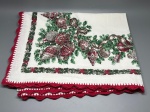 CAMA - MESA - BANHO - Toalha de mesa natalina (200x160 cm) em tecido claro com estampa temática de festas, destacando guirlandas de ramos de pinheiro, fitas vermelhas e bolas e enfeites natalinos ricamente decorados. A peça é arrematada com delicada barra em crochê vermelho trabalhado em pontas ao longo de toda a borda.