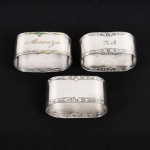 METAL PRATEADO, três (3) porta guardanapos distintos: 1 ornado com guirlandas de tulipas e frisos, medindo 5 x 2,5 x 2,5 cm; 1 ornado com rosas e frisos, marca WOLFF (tradição desde 1894), marca possessória gravada A.J., medindo 4,5 x 2,5 x 2,5 cm; 1 ornado com frisos, marca possessória gravada Mariza, punções de marca não identificado, medindo 5 x 2,5 x 2 cm, vide foto.