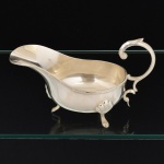 METAL PRATEADO, uma (1) molheira com alça curvilínea terminando em voluta, pés em concha terminando em sapata, borda reforçada em frisos intercalados por X, padrão Império Napoleônico, medindo 20 cm comprimento, sem marca de manufatura aparente, vide foto.