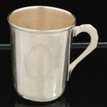 METAL PRATEADO, uma (1) pequena caneca com alças, formato ligeiramente obcônico, ornada com escudo, sem marca de manufatura aparente, possui pequenas mossas, vide foto.