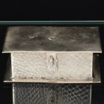 METAL PRATEADO, uma (1) antiga caixa retangular, tampa e fundo lisos, com sinais de que já houve algum ornamento encimando a tampa, laterais com texturização de serpente, parte de traz com acabamentos relevados em mascarões, perolados e elementos vegetalistas, fecho geometrizado, sem marca de manufatura aparente, medindo 12 x 8 x 4 cm, vide foto.