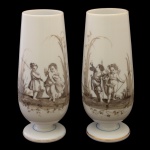 Belíssimo par de vasos floreiros de formato alongado, em opalina leitosa, Apresenta pintura em grisaille e esmalte, retratando querubins e cenas galantes com figuras infantis em trajes de época. Base circular de pedestal adornada com filetes em ouro e tons sutis de azul. 26 cm.