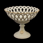 LIMOGES  Fruteira circular funda de pé alto em porcelana francesa branca realçada por detalhes e filetes a ouro. Recipiente vazado por treliças onduladas, apoiado em anelado sobre base circular cônica ornada por monograma. Marcas da manufatura L. Grumbach e da casa comercial em São Paulo sob a base.