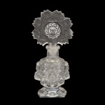 Perfumeiro em cristal transparente. Bojo cilíndrico, ornamentado por máscaras e adornos em alto relevo. Base chanfrada e esparramada lisa. Tampa arqueada, bordada por estrelas centralizando medalhões com motivos raiados. 17 x 8 cm