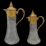 Conjunto de 2 wine decanter em cristal com base bojuda encimada por longo gargalo reto, lapidado e ornado por estilizações geométricas. Tampa e alças em metal dourado. 38 x 18 cm e 30 x 15 cm.