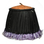 CHAPÉU JUJU  Povo Bamileque. Indumentária de parede feito com penas naturais da África, conhecido como Juju Hat. Apresenta formato levemente cônico, construído com fitas em tecido preto rematadas por plumas roxas. Topo circular em palha trançada rematado por pequena pega.30 x 48 cm (fechado). 14 x 70 cm (aberto). Acervo Particular MAVP.