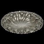 Salva em prata de lei repuxada e cinzelada, com borda recortada e lobulada, e margem decorada por volutas intercaladas por florões. Centro moldado com gomos torcidos, cinzelados e ornamentados por adornos florais. Sob a base contraste do teor de 800 mls. e marcas não registradas. 5 x 30 x 24 cm. Peso 421 gramas. Acervo Particular Maria Anna do Valle Pereira, SP.