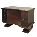 Elegante cômoda Art Déco em madeira nobre, com tampo e caixa de formato retangular com amplo recorte arqueado à frente. Apresenta 3 gavetões superpostos, rematados na base por frisos canelados e salientes, apoiados em largos suportes laterais alongados. Circa 1930-1940. 80 x 117 x 53 cm.  Acervo Particular Maria Anna do Valle Pereira, SP.