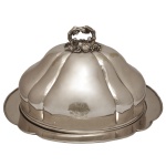 OLD SHEFFIELD  Dish and Food Dome, de formato circular, em metal prateado inglês. Travessa com borda recortada e realçada por debrum. Tampa abobada e moldada em largos gomos, ornamentada por brasão de armas gravado à frente. Pega montada com cachos de uva e folhas de parreira, encimada por argola com galhos entrecruzados. Sob a base, marcas do fabricante E & C. 27 x 50 x 42 cm. Acervo Particular MAVP.