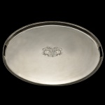 Belíssima bandeja oval em metal espessurado a prata inglês, encimada por galeria ornada por pequenas estilizações repetidas, interrompida por alças vazadas. Centro liso, ornamentado por brasão de armas gravado, apoiado sobre fundo de madeira. Este design clássico e minimalista é característico das peças inglesas. 5 x 80 x 58 cm. Acervo Particular MAVP.