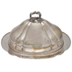 OLD SHEFFIELD Dish and Food Dome, de formato circular, em metal prateado inglês. Travessa com borda recortada e realçada por debrum. Tampa abobada e moldada em largos gomos, ornamentada por brasão de armas gravado à frente. Pega montada com cachos de uva e folhas de parreira, encimada por argola com galhos entrecruzados. Sob a base, marcas do fabricante E & C. Desgastes da prata devido ao uso e tempo. 26 x 45 x 37 cm. Acervo Particular MAVP.
