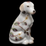 Estatueta em porcelana policromada na forma de um cão sentado. Corpo apresentando decoração floral sobre fundo branco. 15 x 8 x 7 cm.