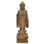 Estatueta em madeira laqueada e dourada, com figura de Budha em pé. Apresenta-se, em atitude de Varada Mudra, tendo a mão para baixo, símbolo de generosidade e compaixão, juntamente em posição de Abhaya Mudra, com a mão para cima, símbolo de destemor e proteção. Juntos, expressam coragem espiritual. Base estilizada em flor de lotos sobre peanha quadrangular com degrau. Sudeste asiático. 62 x 18 cm (MT). 11 x 18 x 15 cm (base_.