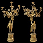 Par de candelabros em bronze ormolu construídos com divindades romanas segurando longas cornucópias encimadas por numerosos ramos floridos. Apresentam-se de pé sobre colunas caneladas e bases quadrangulares chanfradas. Pertenceram ao acervo do Jockey Club de São Paulo. 84 x 38 cm / 86 x 38 cm. Base 16 cm.85 x 43 x 15 cm.