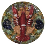 Medalhão decorativo em cerâmica portuguesa, da região de Caldas da Rainha, policromada e esmaltada, apresentando uma lagosta no centro em relevo rodeado por outros mariscos e uma textura que lembra areia. No verso as inscrição: Caldas da Rainha, Portugal. 32 cm.