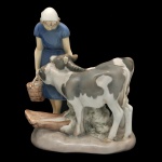ROYAL COPENHAGEN. Bing & Grndahl. Artista Axel Locher. Menina com Bezerro - Estatueta em porcelana dinamarquesa exibindo um acabamento vidrado em tons pastel e suaves, típicos da marca (azul-cinzento, branco e marrom). No verso marca da manufatura Bing & Grndahl, modelo número 2270. 21 x 20 x 10 cm.