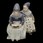 ROYAL COPENHAGEN. Artista Lotte Benter. "Amager Girls Reading" (Garotas de Amager lendo) - Estatueta em porcelana dinamarquesa exibindo um acabamento vidrado em tons pastel e suaves, típicos da marca (azul-cinzento, branco e marrom), retratando duas meninas em trajes tradicionais da ilha dinamarquesa de Amager. No verso marca da manufatura e modelo número 1395. 18 x 17 x 14 cm.