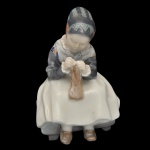 ROYAL COPENHAGEN. Artista Lotte Benter. "Menina de Amager Costurando") - Estatueta em porcelana dinamarquesa exibindo um acabamento vidrado em tons pastel e suaves, típicos da marca (azul-cinzento, branco e marrom). No verso marca da manufatura e modelo número 1314. 16 x 10 x 8 cm.