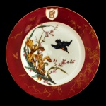Prato contemporâneo em porcelana policromada, com larga borda decorada por insetos e vegetais sobre fundo de cor vinho, apresentando escudo com monograma. Fundo ornado por pássaro e ramos floridos. 23 cm.