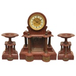 Original e rara Garniture de cheminée, esculpida em jaspe vermelho com colunatas e bases em bronze dourado. Constando de relógio com mostrador branco, montado sobre suporte recortado e ornado por colunatas caneladas nas laterais e na frente com bases cilíndricas ornamentadas por figuras de putti. Acompanham par de fruteiras circulares com pés altos, apoiadas em pedestais construídos no mesmo estilo. Peça de coleção. 47 x 38 x 18 cm (relógio). 27 x 17 x 14 cm (fruteiras).