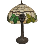 Luminária de mesa estilo Art Nouveau, com coluna em bronze patinado imitando tronco de árvore. Cúpula cônica, montada com placas em vidro marmorizado e policromado, apresentando galho com folhas de parreira e cachos de uva, encimada por ponteira. Base circular. 56 x 40 cm. Apresenta uma placa quebrada.