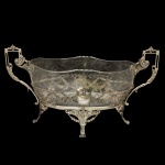 Centro de mesa em prata portuguesa. Formato oval, em cristal e prata repuxada e cinzelada, decorada com guirlandas e folhagens em estilo neoclássico. Bojo com borda ondulada, gravado e decorado com folhagens e estilizações. Suporte vazado e ornado com adornos fitomórficos centralizando escudos, ladeado por alças laterais retas. Base apoiada em pernas recurvas com elementos vegetais, rematadas por pés de garra. Contraste do Porto Javalí 2º título e marca de prateiro desgastada. 28 x 51 x 18 cm (MT). PESO TOTAL1K.219 gramas. Acompanha maças verdes em cerâmica. 8 x 8 cm.