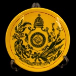 Prato em louça inglesa ou nacional. Fundo amarelo com Brasão de Armas do Império do Brasil no centro encimado pela coroa imperial e rodeado por ramos de café e tabaco. 20 cm. Peça de serviço semelhante ao reproduzido no Livro A Louça da Aristocracia no Brasil, de Jenny Dreyfus à pág. 203. Coleção Antônio Villares da Silva. Apresenta bicado no parte de traz que não compromete a frente. Coleção D. Lucia e Dr. Armando de Arruda Camargo.