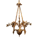 Lustre em bronze dourado de inspiração naturalística. Apresenta cesta vazada, sustentando braços no formato de galhos com folhas, rematados por bobeches. Hastes laterais retas e reunidas ao topo por fita com laço. Acompanha canopla e corrente. 100 x 60 cm.