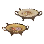 Par de centros de mesa em porcelana pintada à mão. Apresentam decorações florais policromadas com detalhes em tons suaves. Montagens em bronze patinado, com bordas dentadas, ladeadas por alças em argolas e bases rematadas por pés revirados. 7 x 20 x 11 cm.
